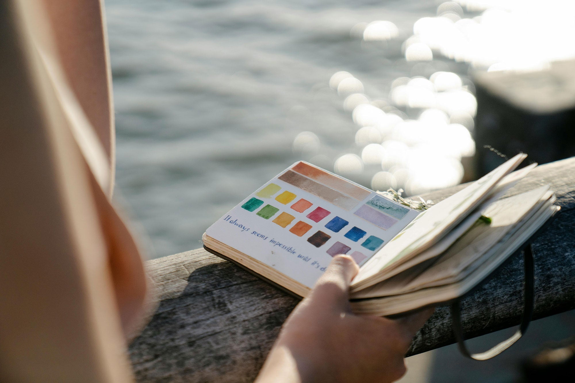 A person holds an open watercolor journal, displaying a vibrant color palette and handwritten note, while sitting by the water with sunlight reflecting on the surface. The scene evokes creativity, mindfulness, and artistic inspiration.