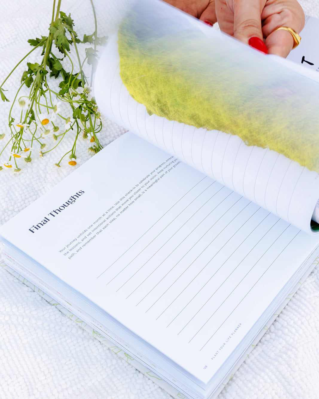 A close-up of an open planner turned to the "Final Thoughts" page, featuring lined sections for reflection and journaling. A hand with red nail polish flips through the pages, with chamomile flowers resting nearby, symbolizing closure, growth, and mindful intention-setting.