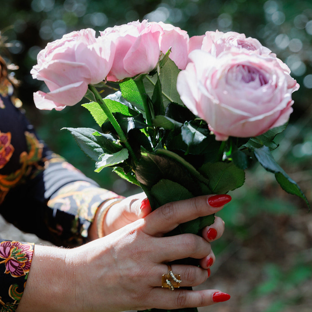 A close-up of a woman's hands holding a bouquet of pink roses. She wears gold rings and has red nail polish, with soft sunlight filtering through the forest, symbolizing beauty, intention, and connection to nature.
