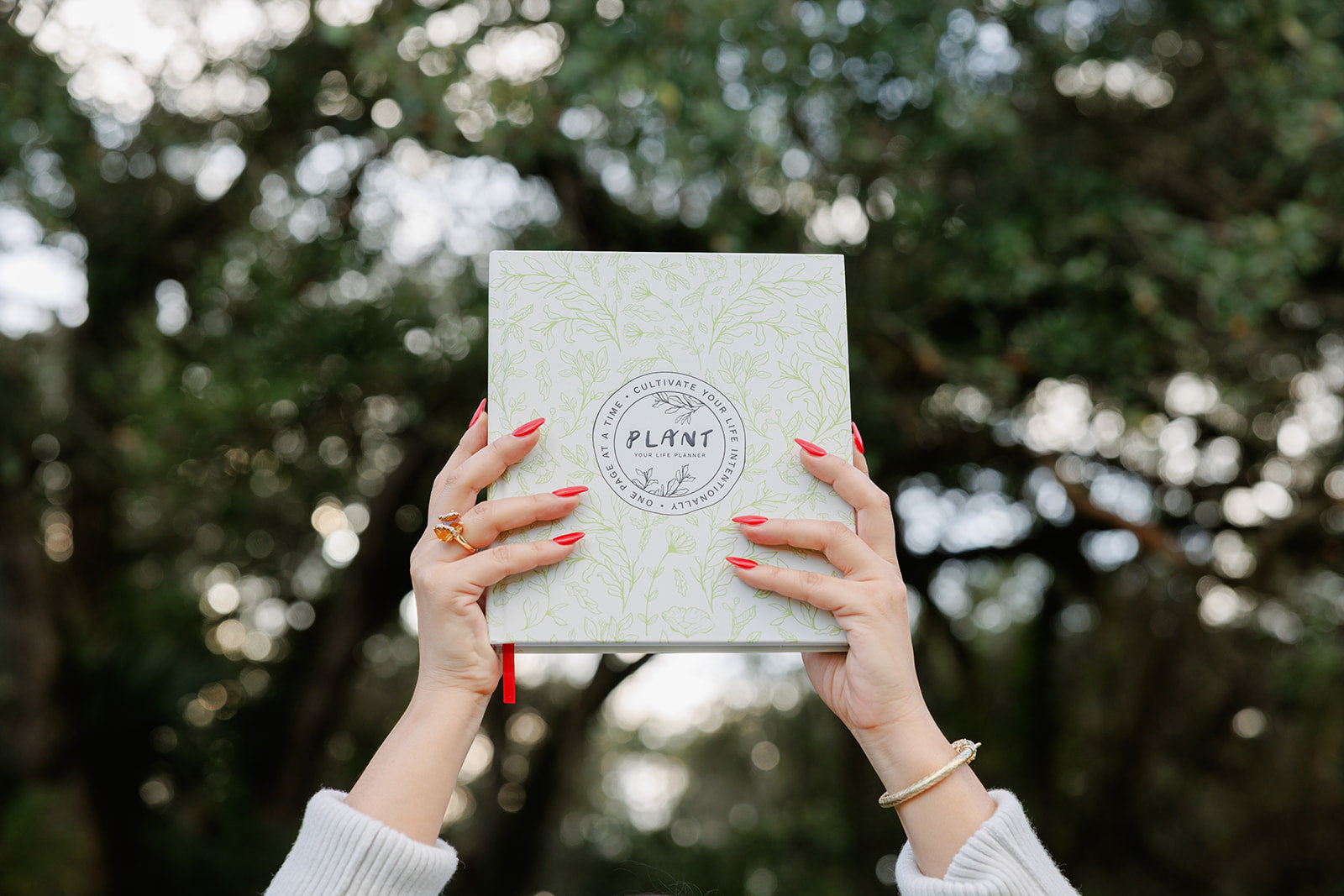 A pair of hands with red nail polish and gold jewelry holds up the "PLANT: Your Life Planner" against a blurred background of lush green trees, symbolizing growth, intention, and new beginnings.