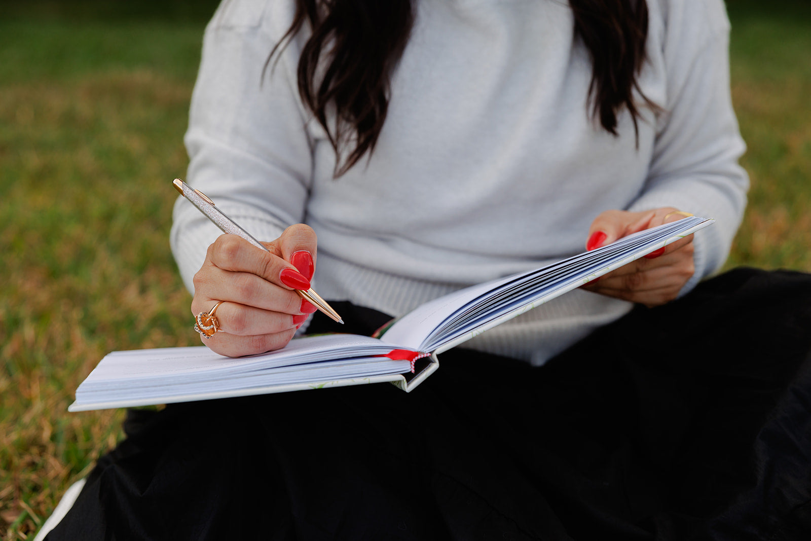A woman sitting on the grass, writing in an open planner with a gold and glitter pen. She wears a white sweater, black skirt, and has red nail polish and gold rings, symbolizing intentional journaling and goal setting.
