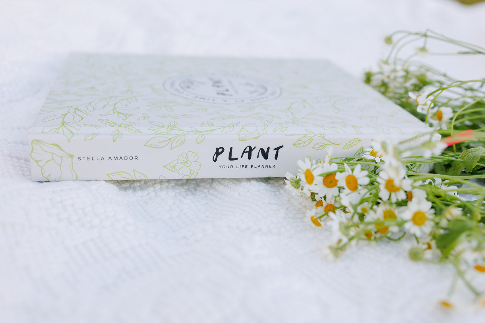 A hardcover planner with a white and green floral design on the cover, titled "PLANT: Your Life Planner" by Stella Amador, resting on a textured white fabric with small white chamomile flowers beside it.