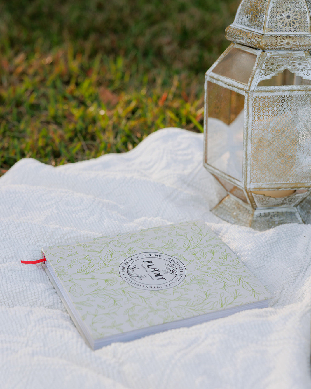 The "PLANT: Your Life Planner" rests on a white textured blanket in a grassy field, accompanied by an ornate white lantern, evoking a peaceful and intentional atmosphere for journaling and reflection.