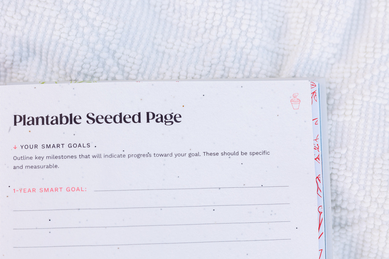 A close-up of an open planner showing a "Plantable Seeded Page" with speckled biodegradable paper designed for goal setting. The page includes lined sections for writing milestones, emphasizing sustainability and intention-setting.
