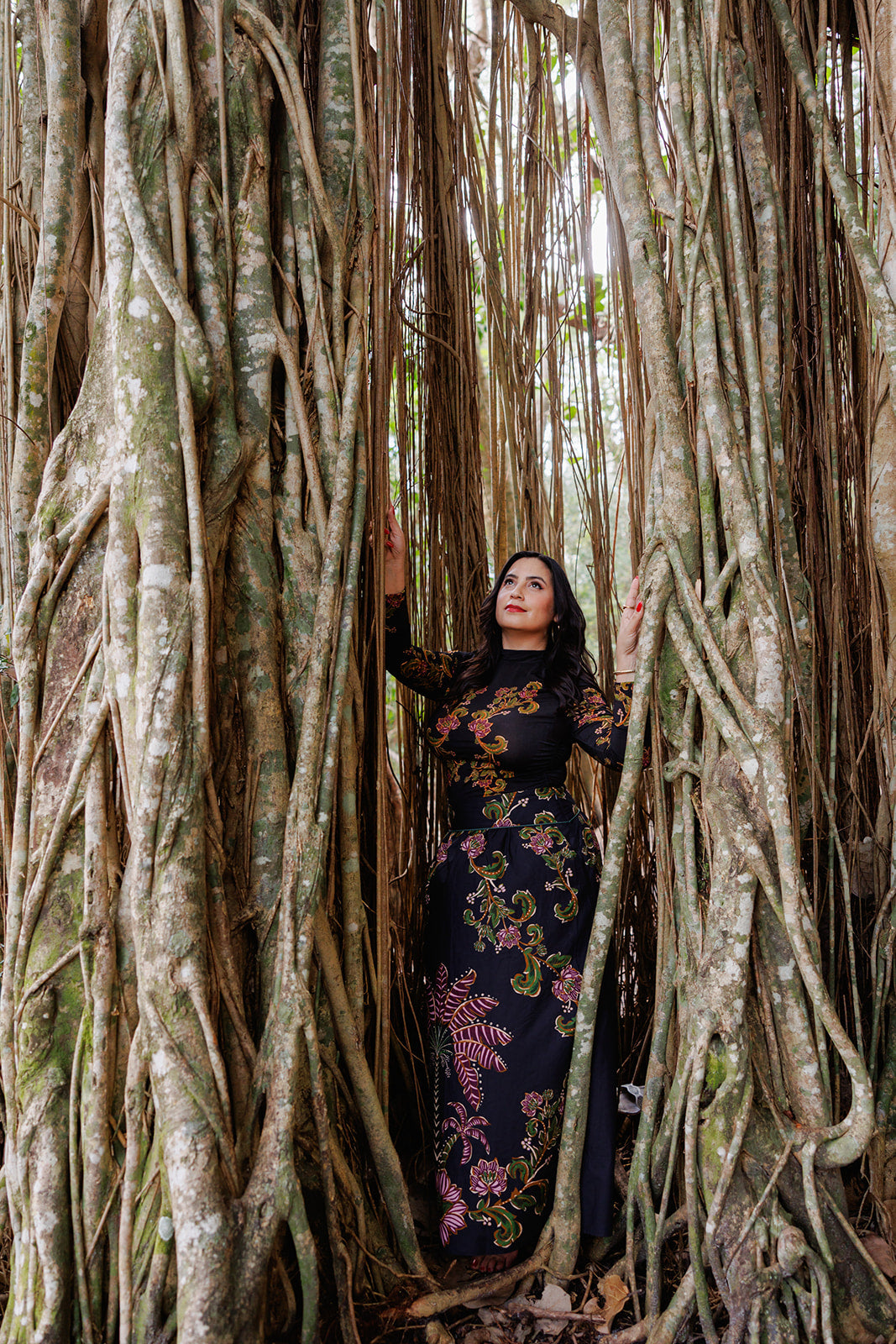 A woman in a black floral dress stands among the towering roots of a banyan tree, gazing upward with her hands touching the vines. The scene evokes themes of strength, connection to nature, and personal growth.