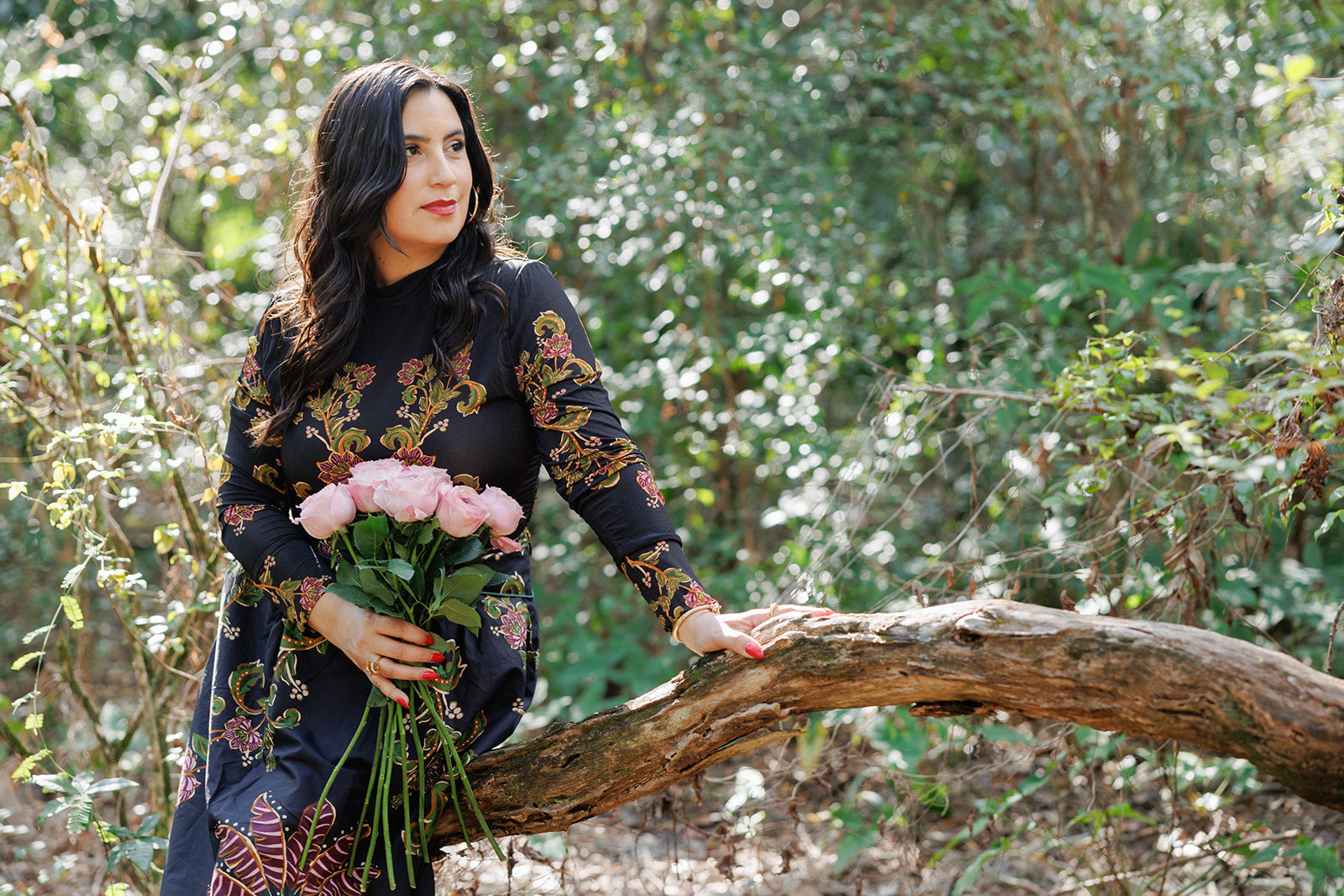 A woman in a black floral dress leans against a fallen tree branch in a sunlit forest, holding a bouquet of pink roses. She gazes softly into the distance, symbolizing tranquility, growth, and connection with nature.