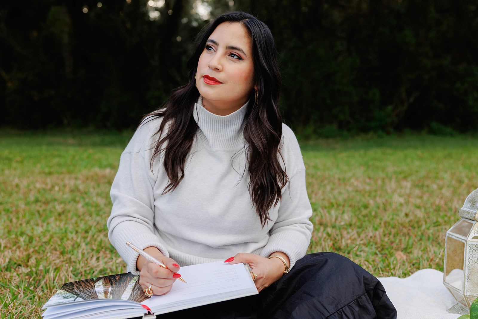 A woman with long dark hair sits on a blanket in a grassy field, holding an open planner and pen. She wears a white turtleneck sweater and black skirt, gazing thoughtfully into the distance, symbolizing reflection and intentional goal setting.