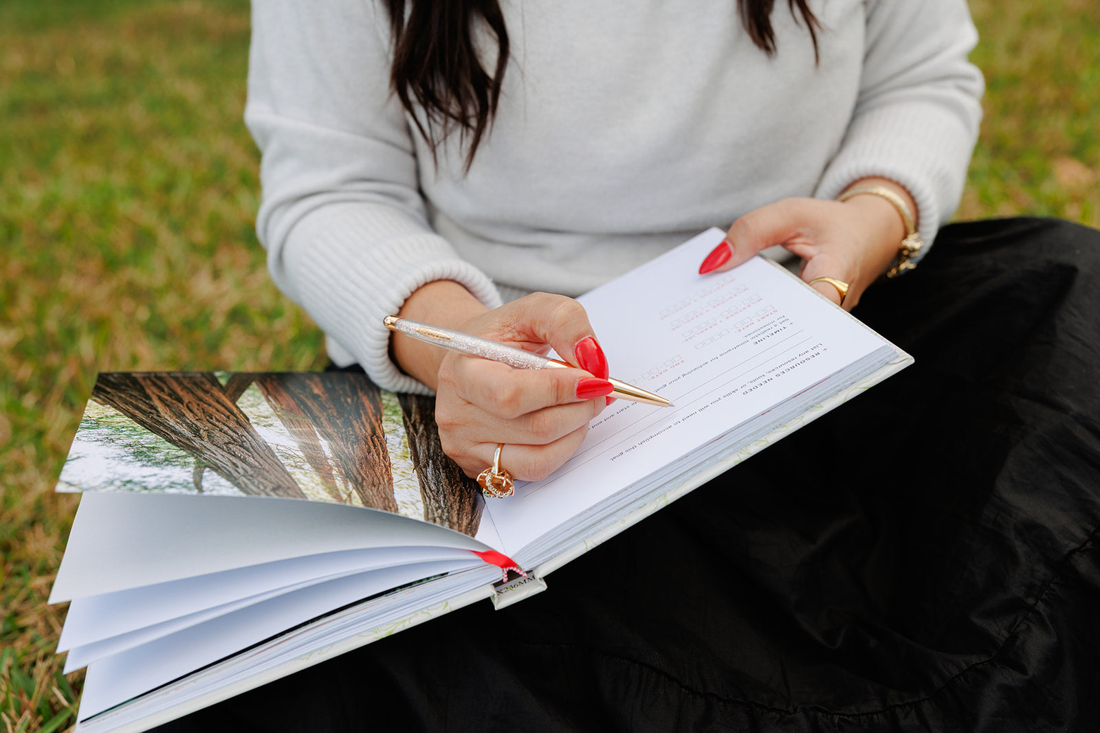 A woman in a white sweater and black skirt writes in an open planner with a gold and glitter pen. She has red nail polish and gold jewelry, with a nature-inspired image visible in the planner, symbolizing reflection and intentional goal setting.